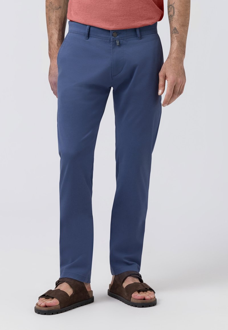 Homme portant un pantalon bleu slim, un t-shirt rouge et des sandales marron avec des boucles, debout devant un fond clair uni.