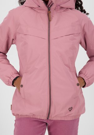 Femme portant une veste à capuche rose clair avec une fermeture éclair à l'avant, des poignets élastiques, des poches avec boutons, et un petit emblème en forme de cœur sur l'ourlet de la jupe.