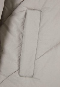 Sección de chaqueta acolchada de color gris claro, con una textura suave y un bolsillo rectangular con costuras limpias en el lado izquierdo.