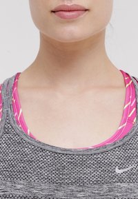 Canotta atletica grigia con un motivo testurizzato e scollatura rotonda, sovrapposta a un reggiseno sportivo a righe rosa e gialle. Logo Nike visibile.