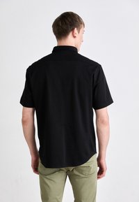 Camisetta nera a maniche corte con orlo dritto e colletto. Tessuto liscio, vestibilità casual, vista da dietro. Abbinata a pantaloni verde chiaro.