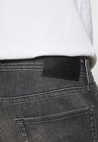 Los jeans de mezclilla gris presentan un parche de cuero negro en la cintura. La tela tiene una textura suave con un ligero efecto de desgaste.