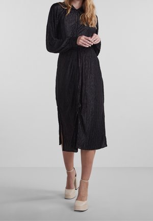 Femme portant une robe noire texturée à manches longues avec une ceinture à la taille et des chaussures à plateforme beige à talons hauts, debout sur un fond uni.