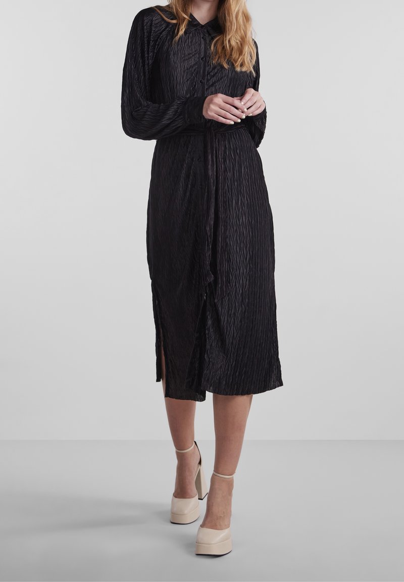 Femme portant une robe noire texturée à manches longues avec une ceinture à la taille et des chaussures à plateforme beige à talons hauts, debout sur un fond uni.