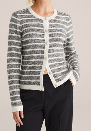 Mannequin portant un cardigan en maille à rayures grises et blanches, avec des bordures blanches et des boutons, associé à un pantalon noir.