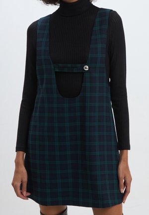 Femme portant une robe salopette à carreaux vert foncé et bleu marine avec un détail de bouton argenté à manches longues sur un pull col roulé noir côtelé.