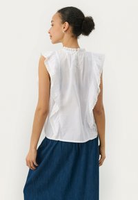 Hvid ærmeløs bluse med volangspidskant og knapdetalje bagpå, parret med en mørk denimnederdel. Blødt stof, afslappet pasform.