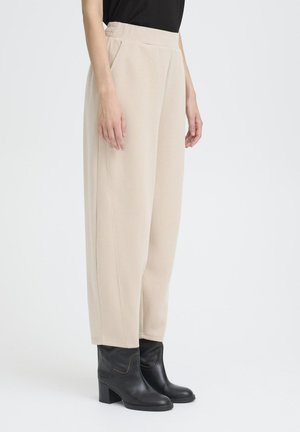 Beige, locker sitzende Hose mit strukturierter Oberfläche, ausgestattet mit seitlichen Taschen und einem hohen Bund, kombiniert mit schwarzen Ankle Boots.