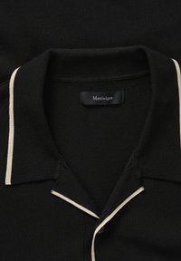 Polo noir avec bordure blanche sur le col et la patte de boutonnage, avec un col replié et une étiquette de la marque "Matinique" à l'intérieur.