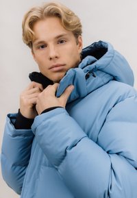 Lyseblå dunjakke med høy krage, med svart innside og glidelås i halsen. Dunstekstur og elastiske mansjetter.