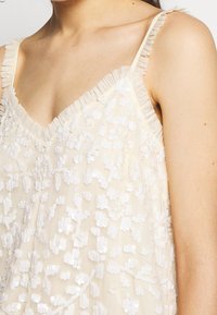 Top sans manches beige avec décolleté en V, présentant un tissu transparent et des motifs floraux en sequins blancs. Les bords ont des accents volantés pour ajouter de la texture.