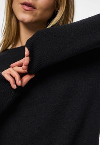 Közeli kép egy személyről, aki egy fekete kötött pulóver ujját igazítja, látható a szövet textúrája és az alsó arc részlete.