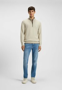 Light beige knitted sweater with a zip collar, paired with light blue jeans and white sneakers. Minimalist logo detail on the chest.