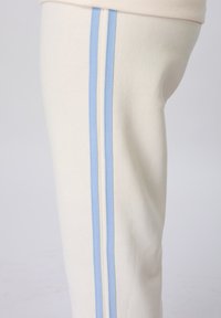 Gros plan sur un pantalon blanc avec deux rayures verticales bleu clair courant le long de la couture latérale sur un fond uni.