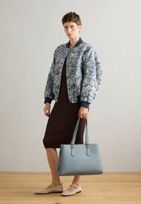 Gėlėtas bomber tipo švarkas mėlynos ir baltos spalvos, briaunotas rudas suknelė, šviesiai mėlyna rankinė su dviem rankenomis ir bežinės spalvos baleto bateliai. Medinė grindų danga.