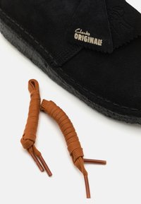 Pantof cu piele de suede negru, cu o talpă flexibilă neagră și cusături decorative. Include două șireturi texturate maro cu agrafe metalice.