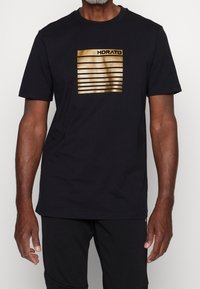 Black cotton t-shirt featuring a gold foil rectangular graphic with horizontal stripes and the word "MORATO" at the top. Short sleeves.