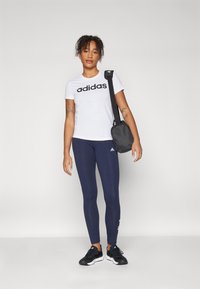 Bílá trička Adidas s černým logem, námořnické legíny a černé sportovní boty. Černá crossbody taška se strapcem přes rameno.