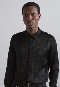 Camicia nera con bottoni, caratterizzata da sottili motivi adonde, texture liscia, maniche lunghe e colletto classico, indossata da un modello.