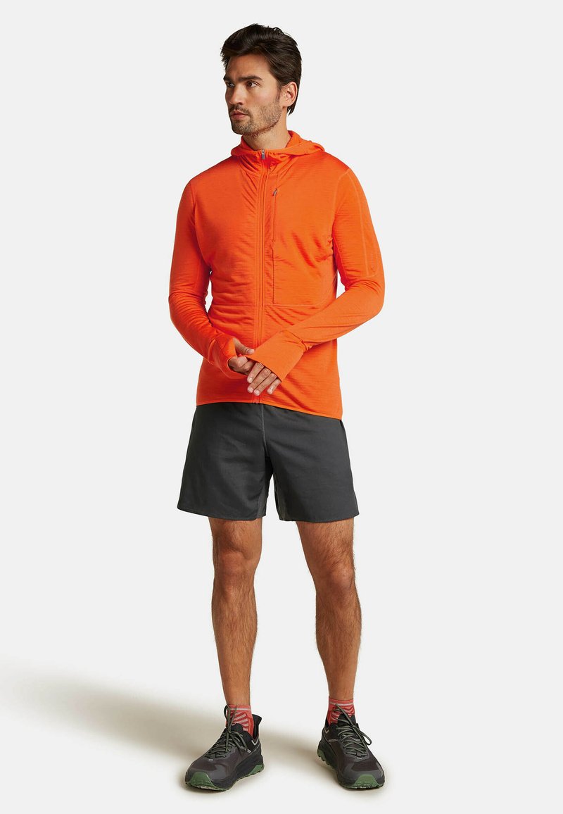 Maglia a maniche lunghe arancione da uomo con cappuccio e zip davanti, abbinata a pantaloni corti scuri e sneakers grigie. Il tessuto sembra leggero e strutturato.