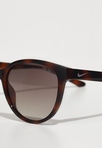 Lunettes de soleil en écaille de tortue brune avec des verres à dégradé sombre. Comprend un épais cadre et un logo Nike discret sur le côté.