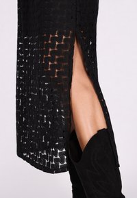 Schwarzes Spitzenkleid mit komplexem Gewebe-Muster und seitlichem Schlitz, kombiniert mit einem schwarzen Wildleder-Stiefeletten, das dekorative Nähte aufweist.