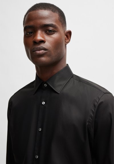 Chemise noire à boutons en tissu lisse, avec un col classique et un léger éclat, ornée de boutons contrastants sur le devant.
