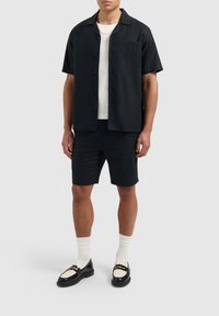 Zwarte kortebloezenshirt met knopen en zwarte shorts, gecombineerd met zwarte loafers met een witte accent en witte sokken. Gladde stofstructuur.