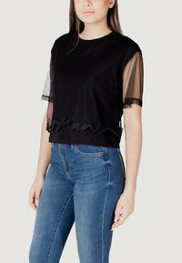 T-shirt nera corta con maniche corte in rete trasparente, caratterizzata da un logo ricamato ondulato sull'orlo. Abbinata a jeans in denim blu.