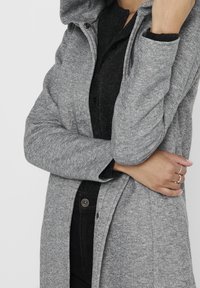 Abrigo gris con capucha y tejido texturizado, con cierre delantero de cremallera y broches. La modelo lleva una camisa negra abotonada debajo.