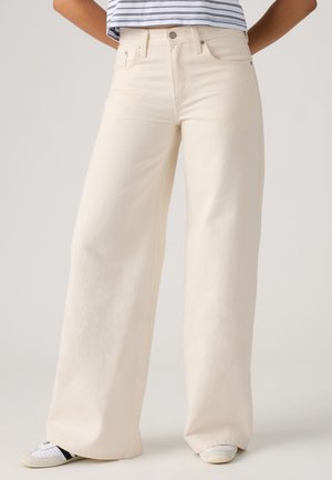 Person iført højtaljede, vidde cremefarvede bukser kombineret med hvide sneakers og en stribet cropped skjorte, stående mod en ensfarvet baggrund.