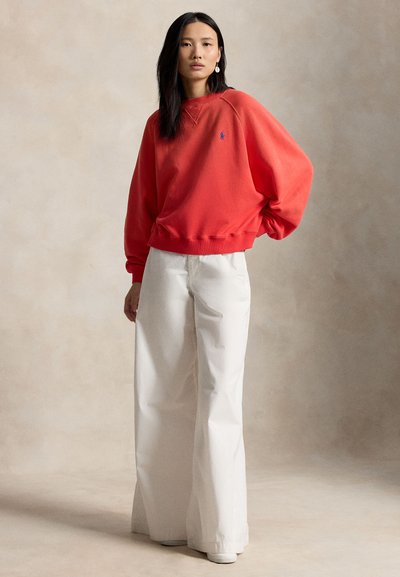 Γυναίκα που στέκεται φορώντας ένα φαρδύ κόκκινο φούτερ με ένα μικρό μπλε λογότυπο και φαρδιά λευκή παντελόνια μπροστά σε μια μονόχρωμη μπεζ φόντο.