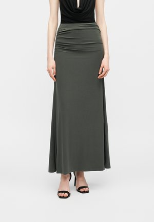 Γυναίκα που φοράει μακριά φούστα σε σκούρο ελιά πράσινο με διακριτική ρυτιδωτή λεπτομέρεια στη μέση, συνδυασμένη με μαύρα ψηλοτάκουνα σανδάλια και μαύρο τοπ.