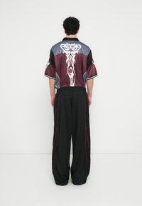 Κοντομάνικο πουκάμισο σε μαροκινό και γκρι με φυλετικά λευκά μοτίβα, συνδυασμένο με φαρδιά μαύρα παντελόνια με κόκκινες λεπτομέρειες και ζώνη με κορδόνι.