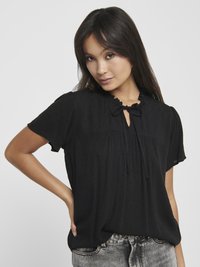 Black short-sleeved blouse with a textured surface, frilled neckline, keyhole detail, and tie closure. Worn with grey high-waisted jeans.