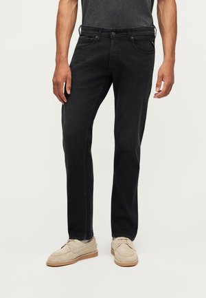 Mand iført slim-fit sorte jeans og beige ruskindssko står mod en ensfarvet lys baggrund.