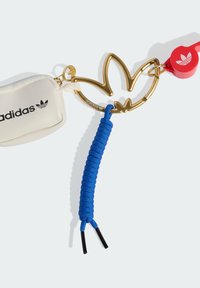 Breloczek z logo metalowym w kolorze złotym w kształcie trzech liści, przymocowany do białego woreczka z czarnym nadrukiem oraz niebieskiego sznurka z czarnymi końcówkami.