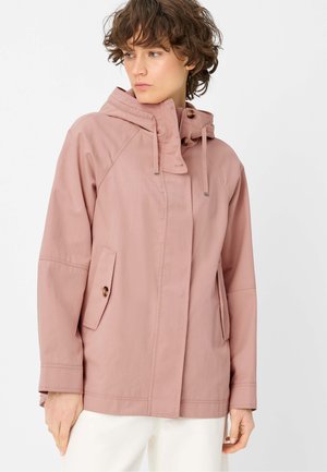 Frau trägt eine staubig rosa Kapuzenjacke mit Knopflaschenklappen und steht vor einem schlichten weißen Hintergrund, blickt nach links.