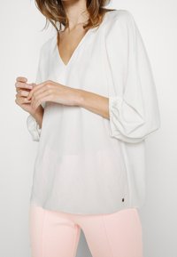 Vit, lätt blus med V-ringning och samlade ärmar med tre fjärdedelar. Har en lätt lyster och en liten gulddetalj vid fållen.