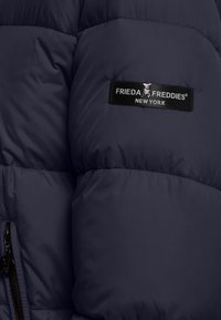 Donkerblauwe pufferjack met een geborduurd textuur, een opvallend zwart label met "FRIEDA FREDDIES NEW YORK," en een zijrits.