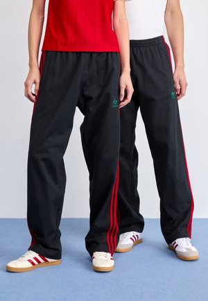 Deux personnes portant des pantalons de survêtement Adidas noirs avec des bandes rouges et des baskets beiges avec des bandes rouges, se tenant sur un sol bleu.