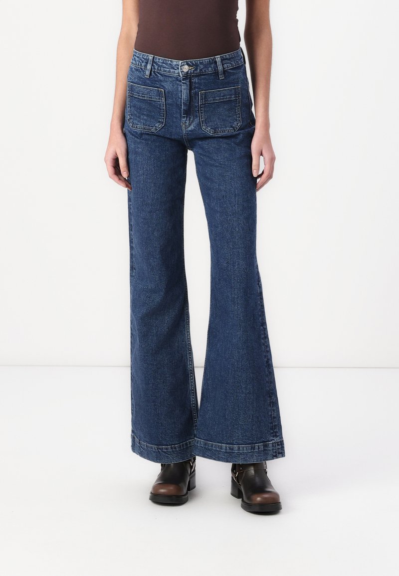 Modre flare jeans z visokim pasom, s tremi zadnjimi žepi in kontrastnim šivom, v kombinaciji z črnimi gležnjarji.