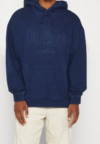 Dunkelblaues Kapuzensweatshirt mit weicher Textur, ausgestattet mit gesticktem "DAP"-Logo und "Harlem" in kontrastierendem Design. Vordertasche vorhanden.