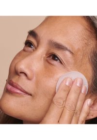 Gros plan sur une personne appliquant un coton sur sa joue. La peau est de ton moyen avec une légère texture, et le coton est blanc et rond.