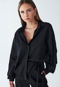 Camicia nera con bottoni realizzata in morbido tessuto, con colletto, maniche lunghe e una vestibilità comoda. Indossata con pantaloni neri abbinati.