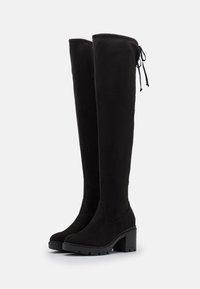 Paire de bottes noires en suède montant jusqu'aux genoux, avec talons épais et semelles crantées, dotées d'un détail lacé au-dessus à l'arrière.