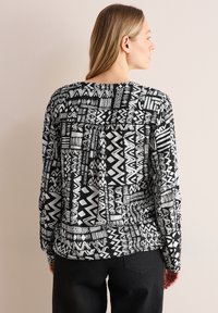 Blouse à motifs noir et blanc avec des designs géométriques, des manches longues, une coupe décontractée et un détail de pli au dos.
