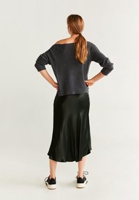 Femme avec une queue de cheval, de dos, portant un pull gris foncé à épaules dénudées, une jupe midi noire et des baskets noires et blanches.