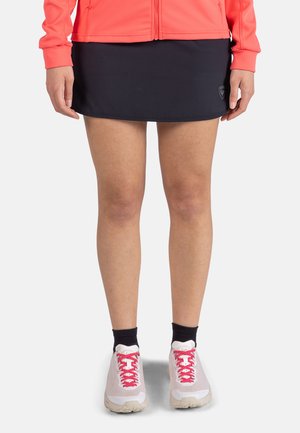 Zwarte sportrok met een gladde textuur, gecombineerd met een koraalkleurige jack met ritssluiting. Witte schoenen met roze accenten en zwarte sokken maken de outfit compleet.