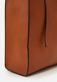 Sac en cuir marron avec une texture lisse, garniture en cuir noir et un design minimaliste ; forme rectangulaire avec des détails de couture visibles.